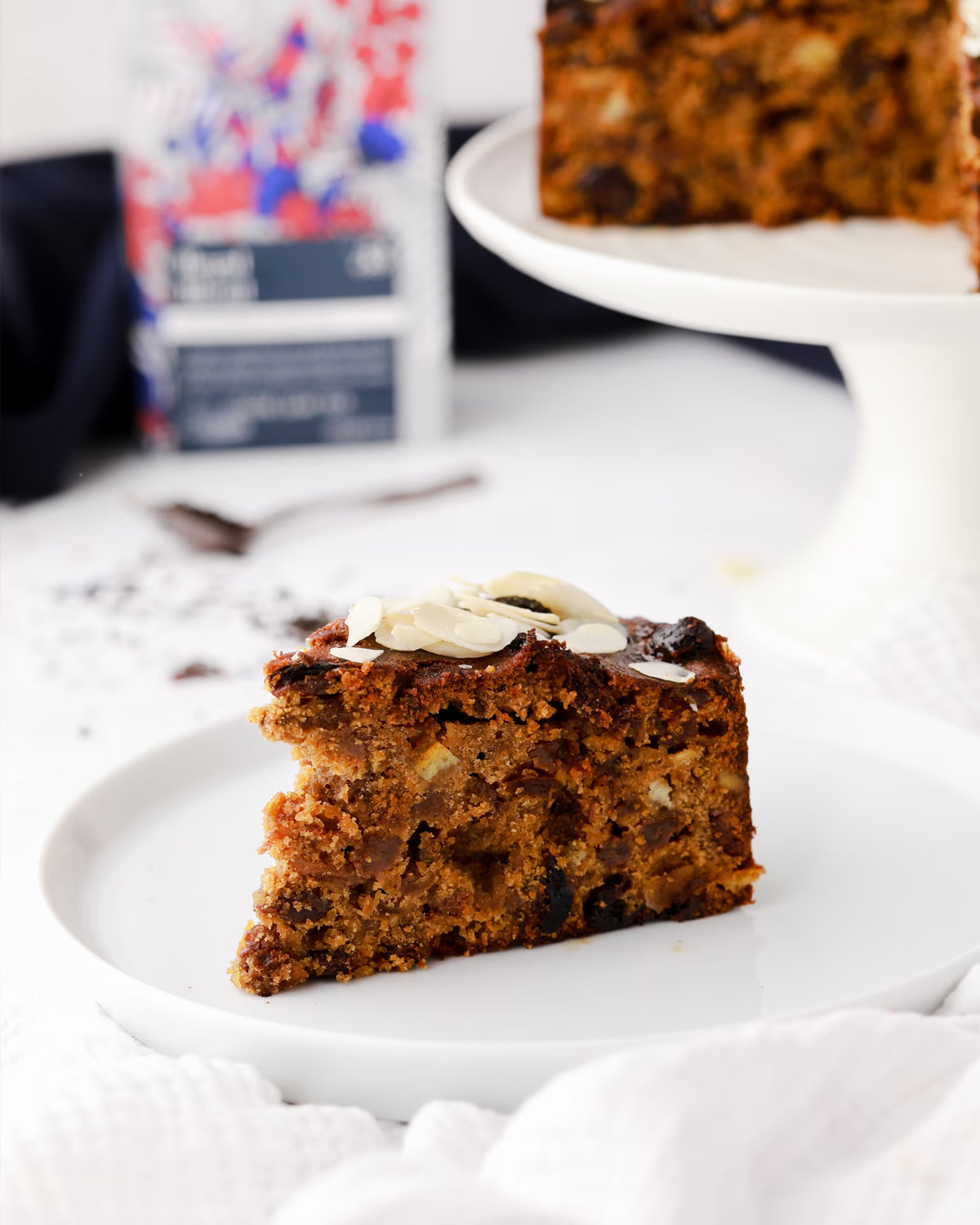 A slice of fruit and nut cake sits on a white plate, topped with almond flakes. The cake is dense with visible pieces of fruit and nuts. A cake stand with another slice of cake is blurred in the background.