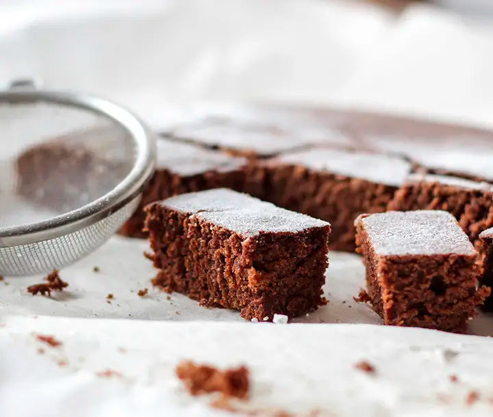 Two pieces of chocolate cake dusted with powdered sugar sit on parchment paper beside a metal sieve, with more cake visible in the background.