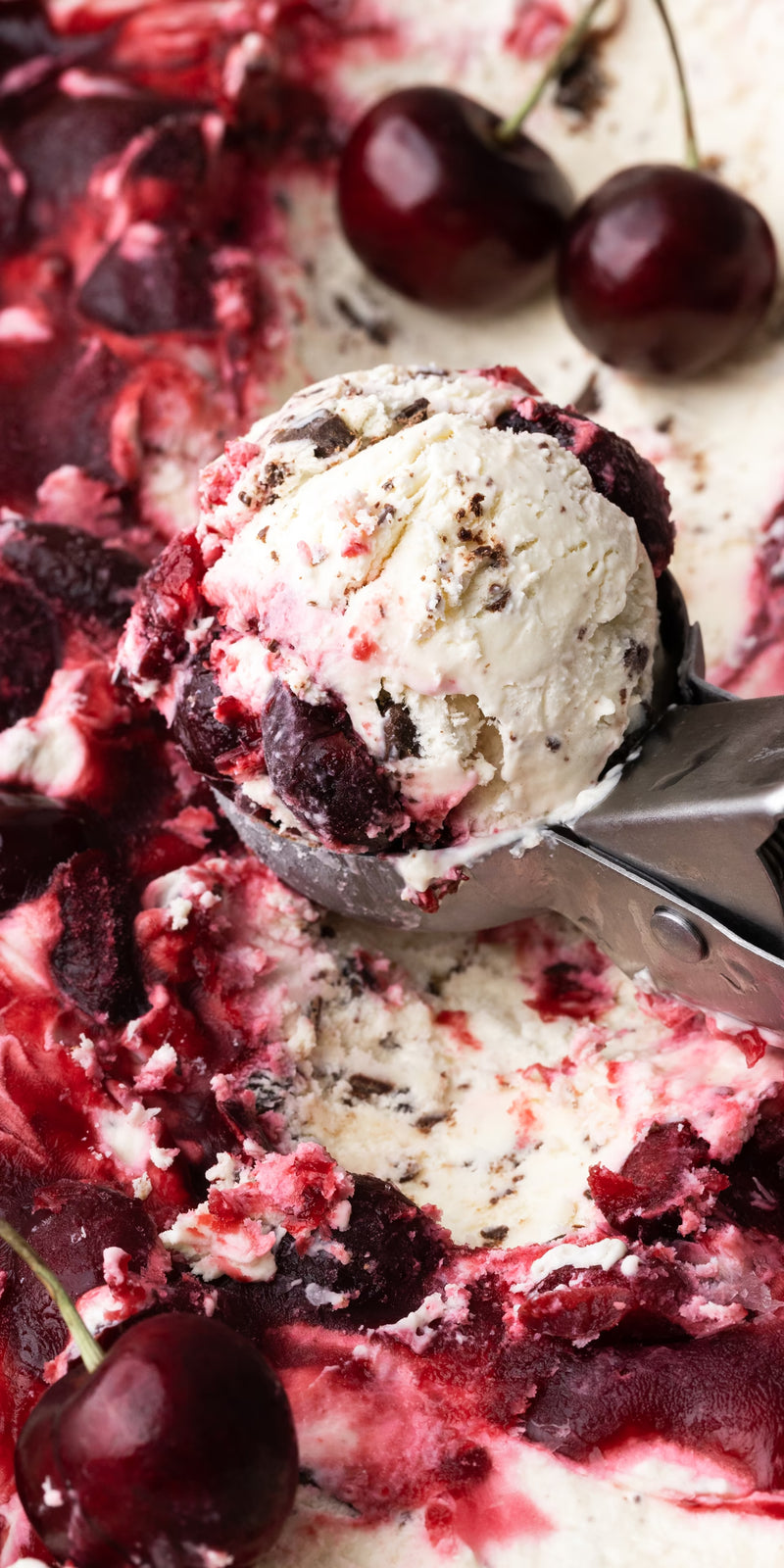 An ice cream scoop lifting cherry and chocolate chip ice cream with swirls of cherry sauce and whole cherries visible, set against a creamy, textured background.