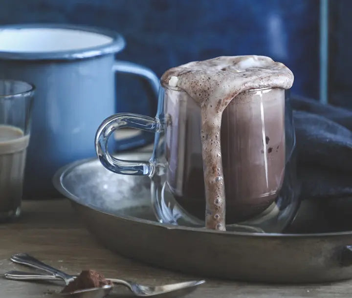 A glass mug filled with frothy hot chocolate sits on a metal tray, with foam spilling over the rim. A blue enamel mug and a glass are in the background, and two spoons rest on the wooden table.