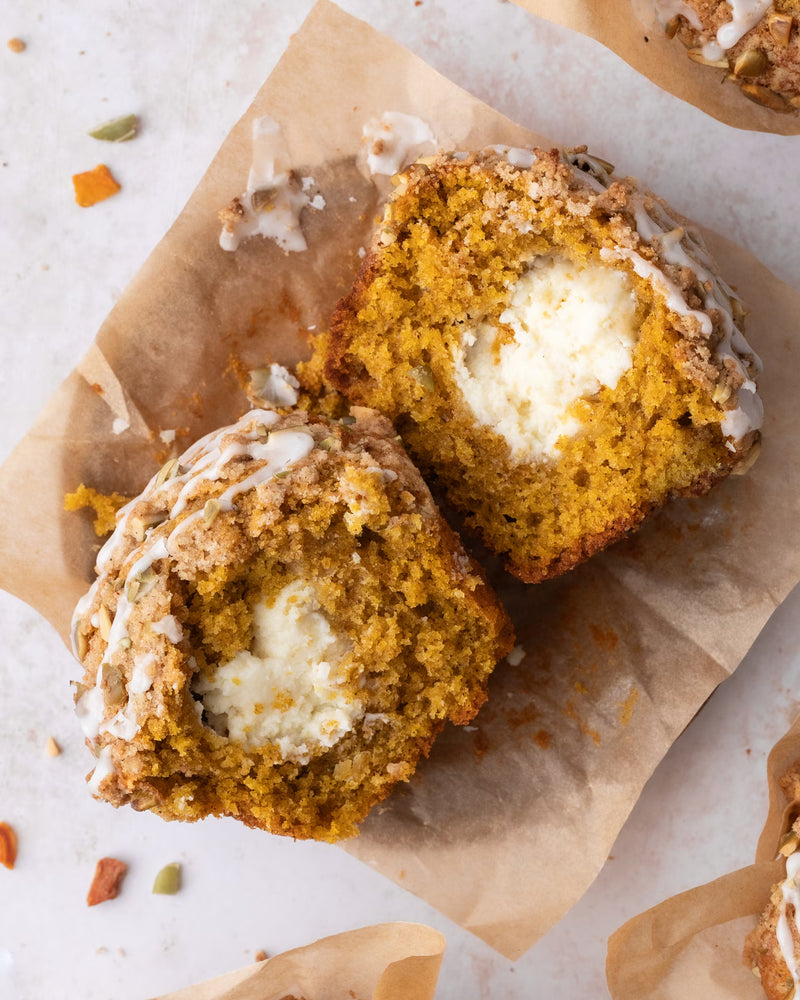 A pumpkin muffin cut in half, revealing a creamy white filling inside. The muffin sits on brown parchment paper and is topped with a light glaze and streusel crumbs.