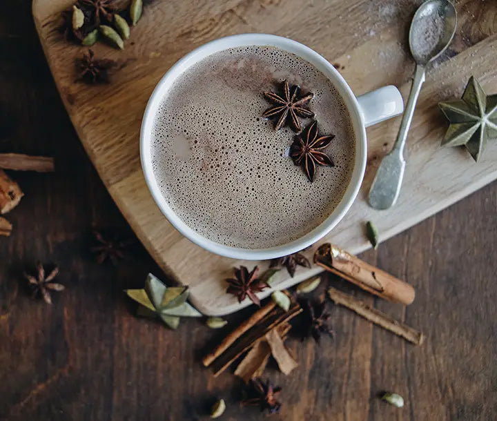 A mug of spiced hot chocolate topped with star anise sits on a wooden board, surrounded by cinnamon sticks, cardamom pods, and a spoon on a dark wood surface.
