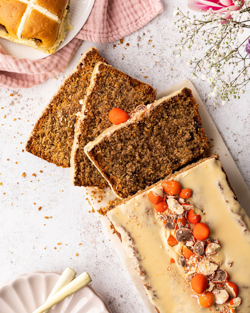 Three slices of carrot cake with icing, garnished with small orange decorations and nuts, sit on a white platter. A partially sliced hot cross bun and flowers are nearby on a textured white surface.