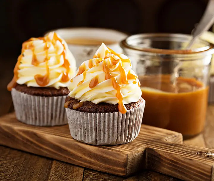 Two chocolate cupcakes with swirled vanilla frosting and caramel drizzle sit on a wooden board, next to a glass jar filled with caramel sauce.