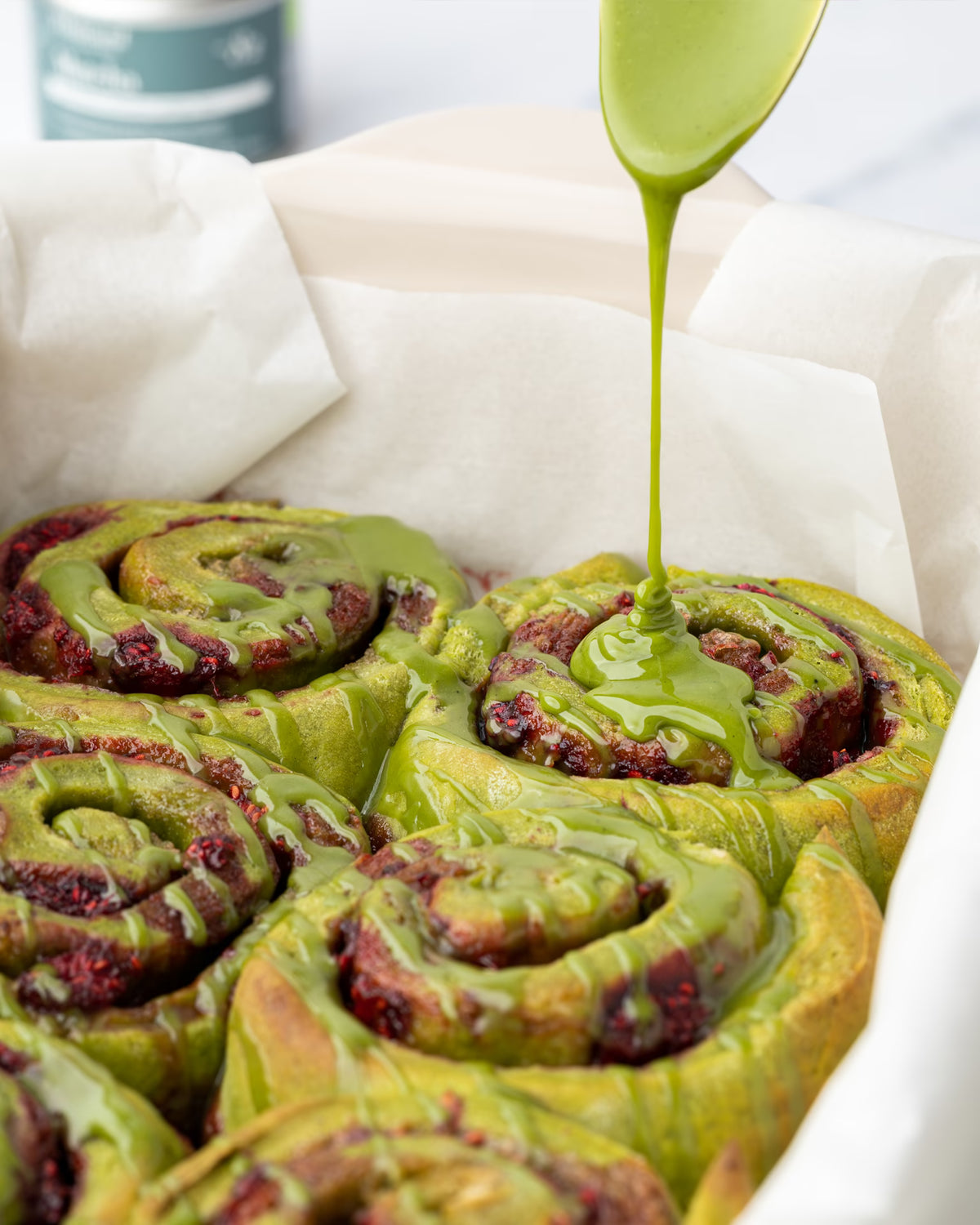 Green icing is being drizzled onto fresh matcha cinnamon rolls with visible berry filling, all nestled in a parchment-lined baking dish.