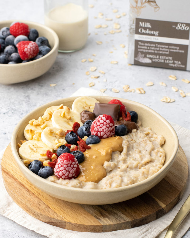 A bowl of oatmeal topped with banana slices, raspberries, blueberries, nut butter, and chocolate sits on a wooden board. A small bowl of berries, a glass of milk, and a tea box are in the background.