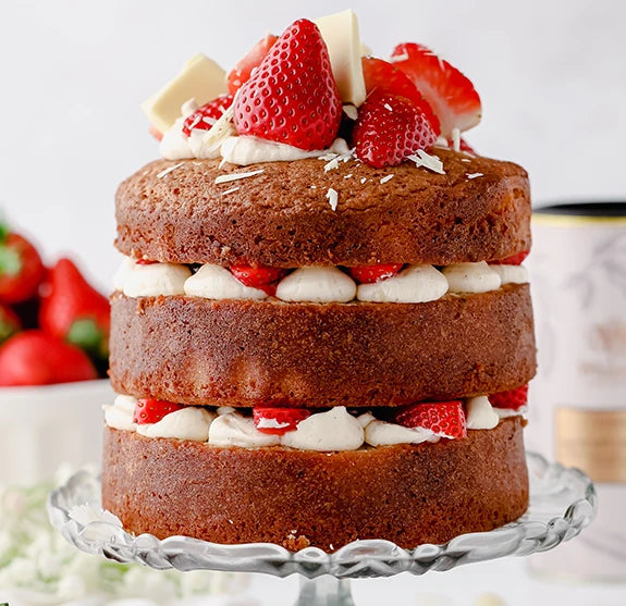 A tall, layered sponge cake with cream and strawberries between layers, topped with fresh strawberries and white chocolate pieces, displayed on a glass cake stand.