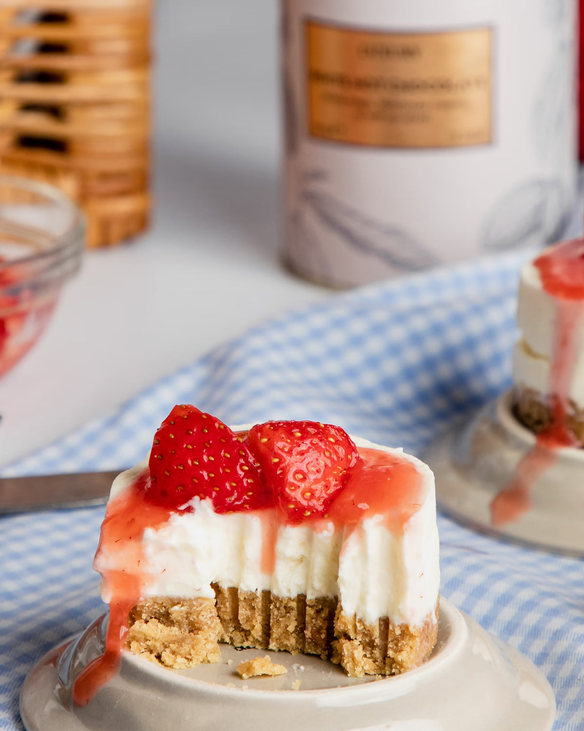 A small cheesecake with a graham cracker crust, topped with strawberry sauce and fresh strawberries, sits on a dish with a bite taken out. A blue checkered cloth is underneath, and blurred items are in the background.