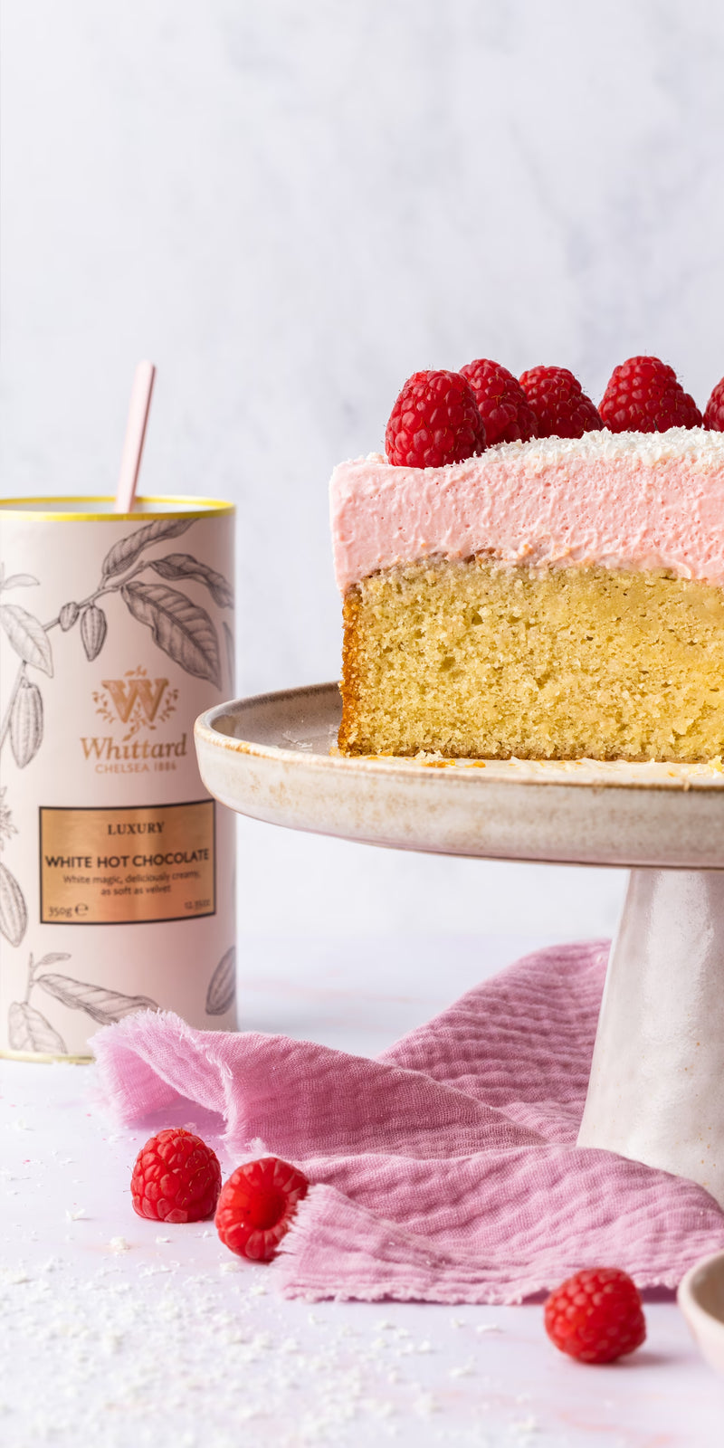 A slice of vanilla cake with pink frosting and raspberries on top sits on a cake stand. A can of Whittard white hot chocolate and loose raspberries are nearby on a pink cloth.